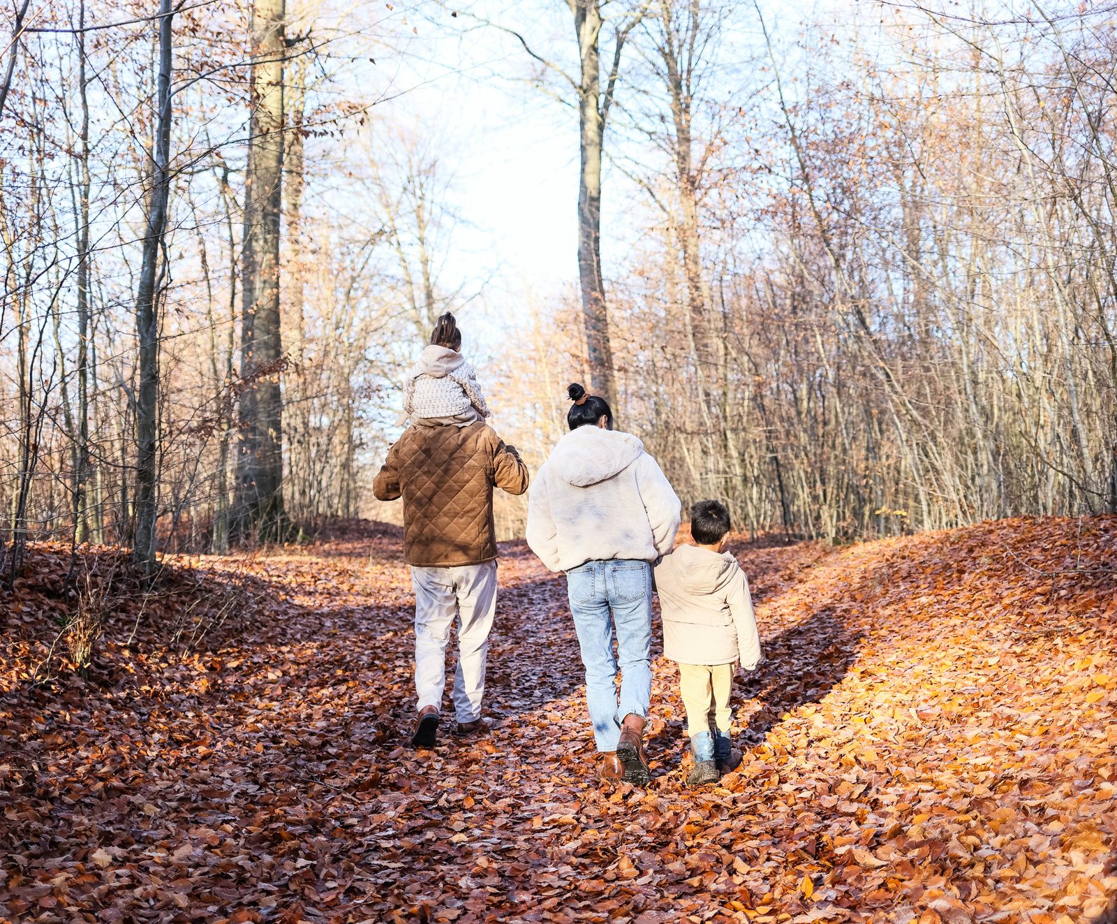 Een gezin van vier wandelt over een bospad vol herfstbladeren; het jongste kind zit op de schouders.