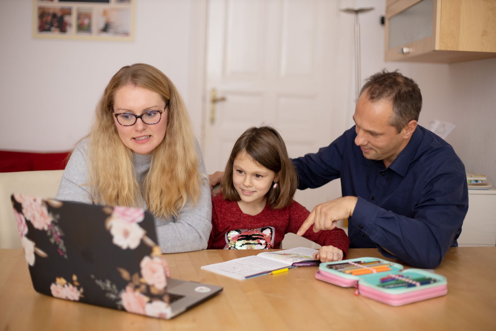 Ouders en hun dochter samen aan de houten tafel, tussen huiswerk, potloden en een laptop.